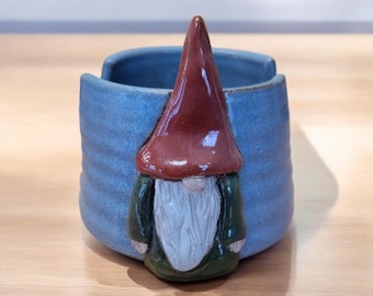 May include: A blue ceramic planter with a gnome design. The gnome has a red hat, a white beard, and a green shirt.