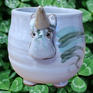 May include: A white ceramic mug with a unicorn face sculpted on the front. The unicorn has a brown horn and green and white painted details. The mug has a rounded shape and a wide base.
