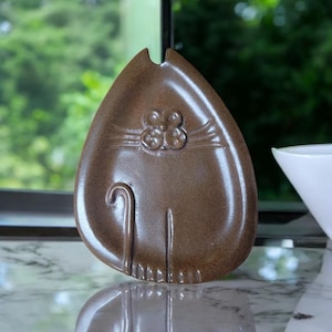May include: A brown ceramic cat-shaped dish with a flower design on its face. The dish is glazed and has a glossy finish.