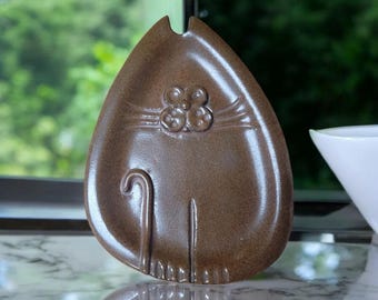 May include: A brown ceramic cat-shaped dish with a flower design on its face. The dish is glazed and has a glossy finish.