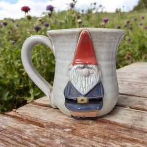 May include: A white ceramic mug with a gnome face sculpted on the front. The gnome has a red hat, a white beard, and a blue jacket. The mug has a handle on the left side.