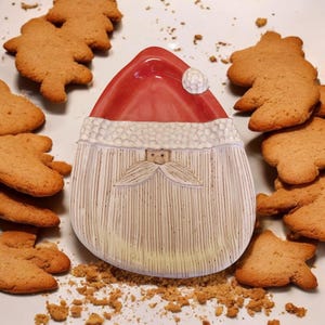 May include: A ceramic Santa Claus dish with a red hat and white beard. The dish is surrounded by gingerbread cookies.