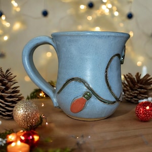 May include: A light blue ceramic mug with a green and brown design of a string of Christmas lights. The mug has a red and orange light bulb on the side.