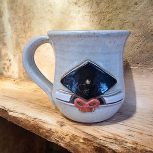 May include: A white ceramic mug with a graduation cap, tassel, and ribbon design. The cap is black with a gold accent. The mug has a handle and is perfect for a graduation gift.