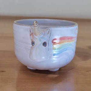 May include: A white ceramic bowl with a unicorn face and a rainbow design. The unicorn has blue eyes and a horn.
