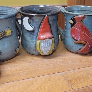 May include: Five handmade ceramic mugs with various designs. One mug features a gnome, another a cardinal, and two have a moon and stars design. The fifth mug is a lidded container with a cardinal design.