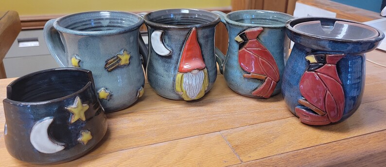 May include: Five handmade ceramic mugs with various designs. One mug features a gnome, another a cardinal, and two have a moon and stars design. The fifth mug is a lidded container with a cardinal design.