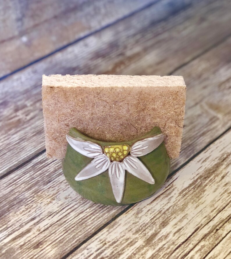 May include: A green ceramic dish with a white flower design. The dish is shaped like a half-circle and has a small opening at the top. The flower has a yellow center and five white petals.