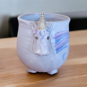 May include: A ceramic mug shaped like a unicorn's head with a white glaze and purple and blue accents. The mug has a horn and a whimsical expression.