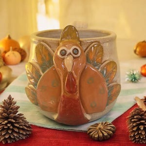May include: A ceramic mug shaped like a turkey with a brown, orange, and white glaze. The turkey has large, round eyes and a red wattle.