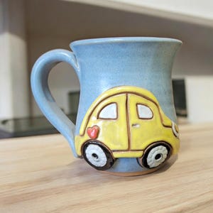 May include: A blue ceramic mug with a yellow car design. The car has a red heart on the side and black wheels.