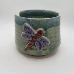 May include: A green ceramic yarn bowl with a dragonfly design. The dragonfly is purple and brown with a textured surface.