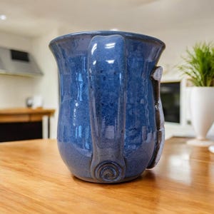 May include: A blue ceramic mug with a unique handle and a white design on the side. The mug is sitting on a wooden table.
