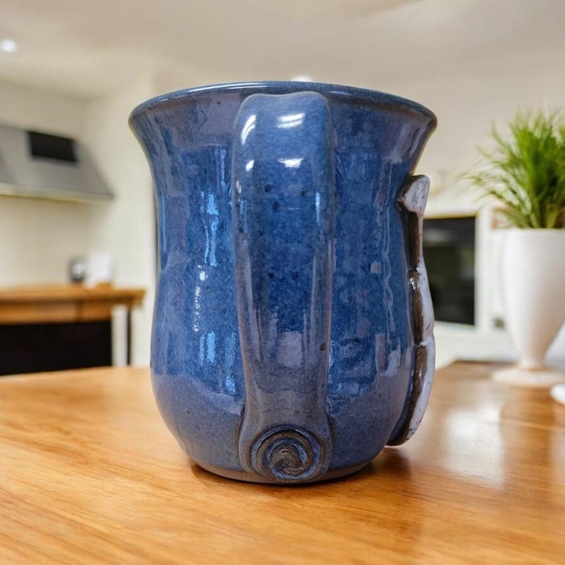 May include: A blue ceramic mug with a unique handle and a white design on the side. The mug is sitting on a wooden table.