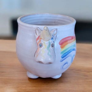 May include: White ceramic mug with a unicorn face and a rainbow painted on the side. The unicorn has a gold horn and blue eyes.