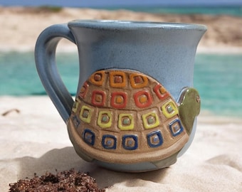 Può includere: Una tazza in ceramica blu con un designo di tartaruga colorato. Il guscio della tartaruga è fatto di piccoli quadrati in tonalità di giallo, arancione, rosso, blu e verde.