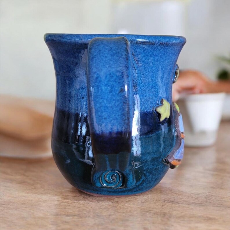 K&ouml;nnte beinhalten: Eine blaue Keramiktasse mit einem fantasievollen Design mit Mond, Sternen und einem Vogel. Die Tasse hat einen strukturierten Griff und eine blaue und gr&uuml;ne Glasur.