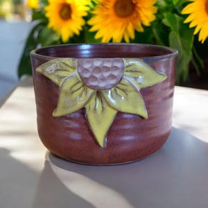 May include: A brown ceramic mug with a yellow and green sunflower design. The mug has a textured surface and is glazed in a brown color.