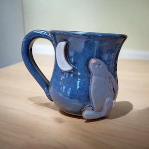 May include: A blue ceramic mug with a white crescent moon and a gray rabbit design. The rabbit is looking up at the moon.