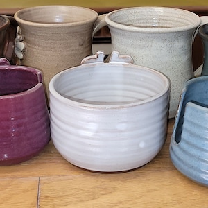 May include: A collection of ten ceramic mugs and bowls in various colors, including purple, white, green, blue, brown, and red. The mugs and bowls have a textured, handmade look.