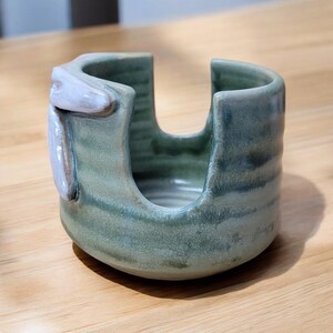 May include: A green ceramic dish with a white glaze, designed to hold a sponge or dish brush. The dish has a unique, organic shape and a textured surface.