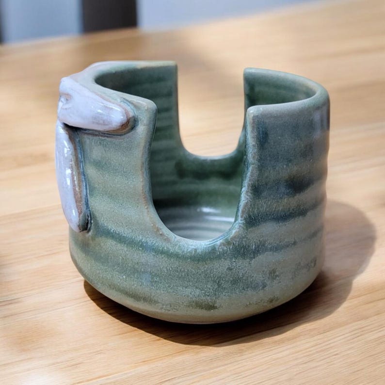 May include: A green ceramic dish with a white glaze, designed to hold a sponge or dish brush. The dish has a unique, organic shape and a textured surface.