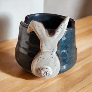 May include: A blue ceramic planter with a white rabbit design. The rabbit has a speckled white glaze and a blue and red glaze on the ears.