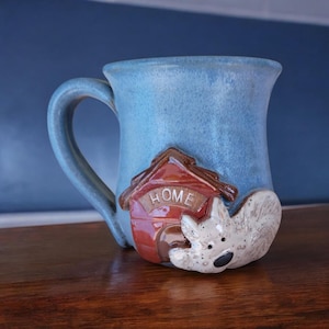 May include: A blue ceramic mug with a 3D dog house and dog design. The dog house is red with the word "HOME" on the front. The dog is white with brown accents.