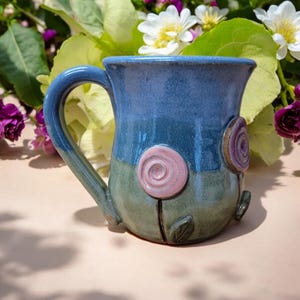 May include: A handmade ceramic mug with a blue and green glaze. The mug has a handle and is decorated with three pink and purple flowers.
