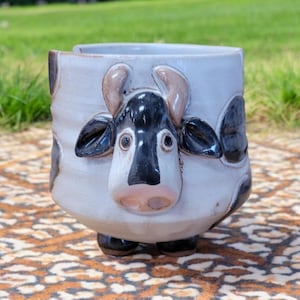 May include: A ceramic cow-shaped mug with black and white spots and a pink nose. The mug has a white glaze and is hand-painted.