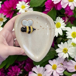 May include: A small, heart-shaped ceramic dish with a bee design. The dish is beige with a light brown glaze and features a yellow and black bee with brown wings. The bee is flying with a dotted line trailing behind it.