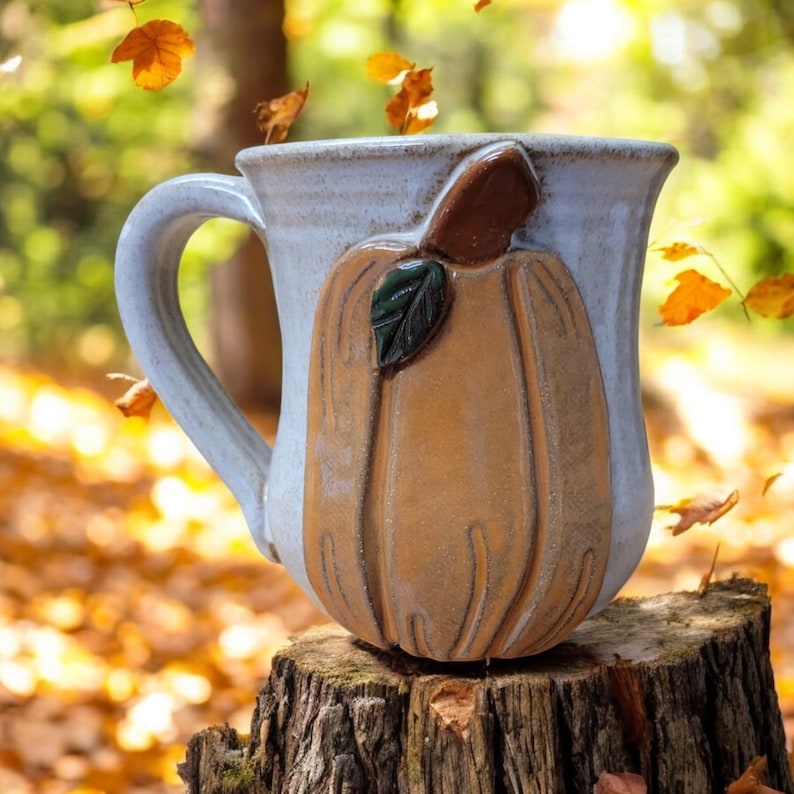May include: A white ceramic mug with a brown and green pumpkin design. The mug has a handle and is sitting on a tree stump.