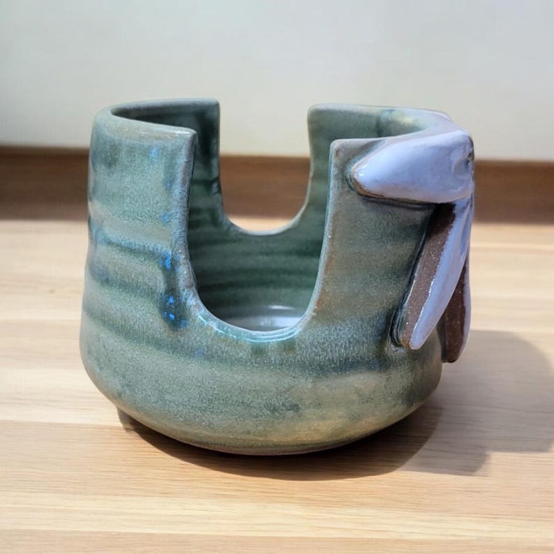 May include: A green ceramic dish with a unique design. The dish has a wide opening and a smaller opening on the side. The dish has a blue and green glaze and a brown and purple design on the side.