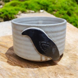 May include: A white ceramic mug with a black crow design. The mug has a textured, slightly speckled surface.