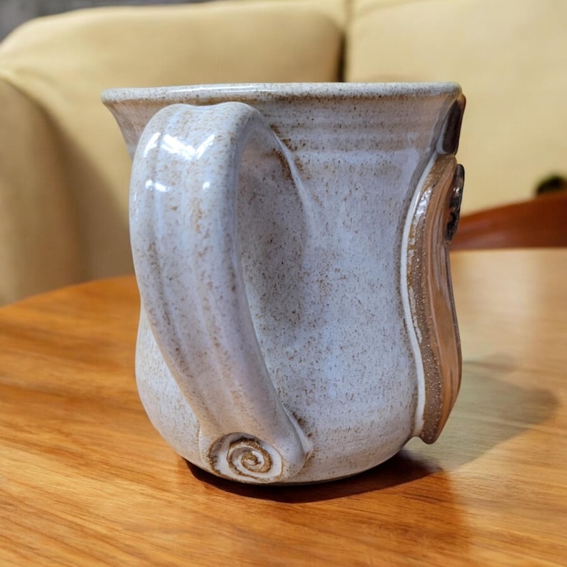 May include: A white ceramic mug with a brown handle and a swirl design on the bottom. The mug has a speckled glaze.