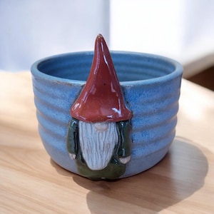 May include: A blue ceramic bowl with a gnome face on the front. The gnome has a red hat, a white beard, and green clothing.