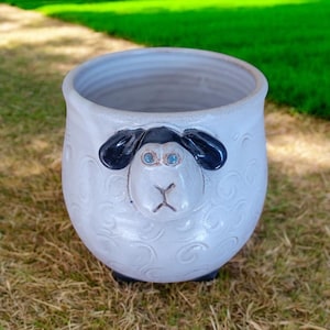 May include: White ceramic planter shaped like a sheep with black ears and blue eyes. The planter has a textured surface that resembles sheep's wool.