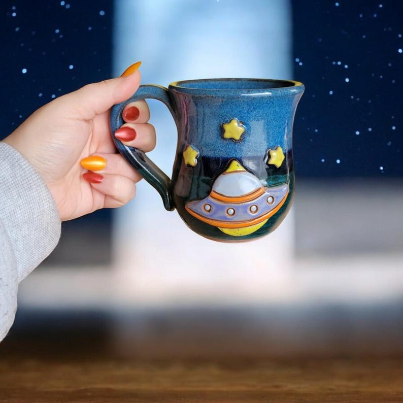K&ouml;nnte beinhalten: Eine blaue Keramiktasse mit einem gelben und orangefarbenen Raumschiffdesign. Die Tasse hat einen Henkel und ist mit Sternen verziert.