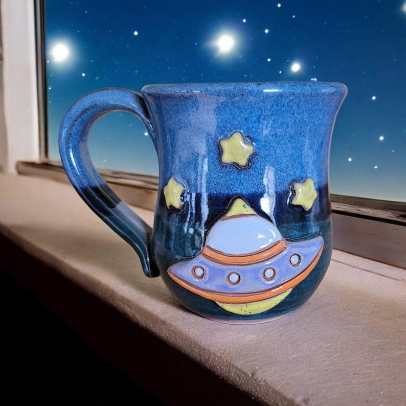 K&ouml;nnte beinhalten: Eine blaue Keramiktasse mit einem Galaxie-Design und einem gelben und violetten Raumschiff auf der Vorderseite. Die Tasse hat einen Henkel und ist mit gelben Sternen verziert.