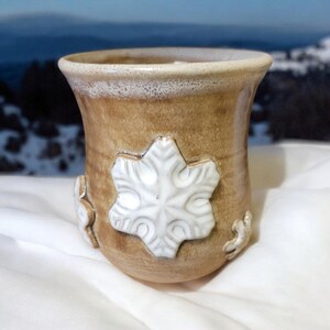 May include: A brown ceramic mug with a white snowflake design. The mug has a rounded bottom and a wide rim.
