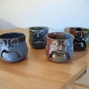 May include: Four ceramic mugs with unique, expressive faces. Each mug is glazed in different shades of blue, green, and brown, with a textured finish. The mugs have a unique design with a cutout for a handle.