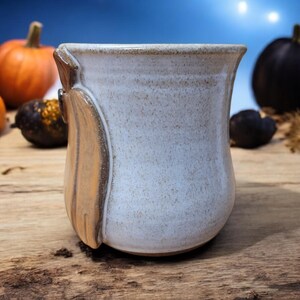 May include: A white ceramic mug with a brown and orange glaze on one side. The mug has a rounded bottom and a wide opening.