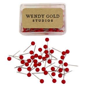 May include: A clear plastic storage box containing red push pins with a tan label printed with the text WENDY GOLD STUDIOS. Below the box, a collection of loose red push pins with silver metal points are scattered across the frame.