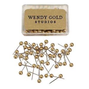 May include: A clear plastic rectangular storage box containing gold-colored ball-head push pins, with a tan label reading WENDY GOLD STUDIOS. A collection of identical gold-colored ball-head push pins is scattered below the storage box.