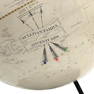 May include: A close-up of a vintage-style globe with the text "SULLIVAN FAMILY ADVENTURES" on a banner. The globe has pins marking locations, with names like "ADAM" and "SUSAN". The globe is beige with black text and a black stand.