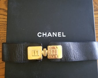 Pode incluir: Um cinto de couro preto com uma fivela dourada com o logotipo da Chanel e as palavras "Coco Chanel". O cinto está em uma caixa preta com a palavra "CHANEL" impressa em branco.
