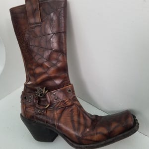 May include: Brown leather cowboy boots with a spiderweb pattern and a metal buckle. The boots have a pointed toe and a block heel.