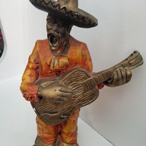 May include: A ceramic figurine of a man wearing a sombrero and playing a guitar. The figurine is painted in shades of orange, yellow, and gold.