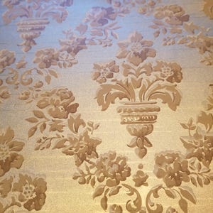 May include: Close-up of a fabric with a repeating floral pattern. The design features bouquets of flowers and scrolling leaves in shades of beige and brown against a light gold background. The fabric has a subtle sheen.