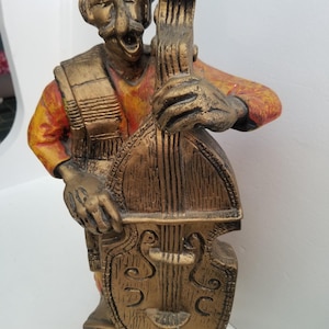 May include: A gold-painted ceramic figurine of a man wearing a sombrero and playing a bass guitar. The figurine is painted in shades of orange, red, and gold.
